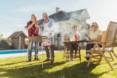 Foto de Dos hombres mayores haciendo un brindis con botellas de cerveza mientras asan carne en el patio trasero con sus esposas sentadas a la mesa en el fondo, tomando café y relajándose. - Imagen libre de derechos