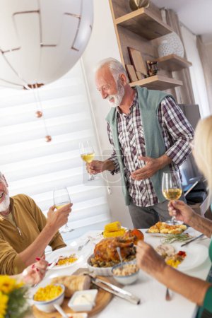 Téléchargez les photos : Des amis aînés se sont réunis autour de la table, célébrant Thanksgiving, dînant ensemble à la maison et levant des verres de vin, portant un toast, accueillant les invités - en image libre de droit