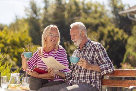 Foto de Feliz pareja de ancianos disfrutando de su tiempo juntos bebiendo café en el patio trasero de su casa, hombre leyendo el periódico mientras que la mujer está leyendo un libro - Imagen libre de derechos