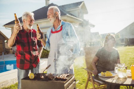 Téléchargez les photos : Hommes âgés qui s'amusent à porter un toast avec des bouteilles de bière et à griller de la viande pendant que les femmes sont assises et se relaxent en arrière-plan ; personnes âgées ayant une fête barbecue arrière-cour - en image libre de droit