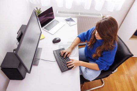Téléchargez les photos : Vue grand angle du codage du programmeur féminin tout en travaillant au bureau à domicile ; développeur Web féminin travaillant à distance à partir de la maison à l'aide d'ordinateurs de bureau et d'ordinateurs portables - en image libre de droit