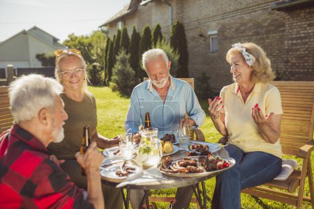 Foto de Grupo de alegres amigos mayores almorzando al aire libre en el patio trasero, reunidos alrededor de la mesa, comiendo, bebiendo y relajándose en un soleado día de verano. - Imagen libre de derechos