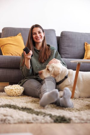 Téléchargez les photos : Belle jeune femme relaxante à la maison, assise sur le sol du salon regardant la télévision et mangeant du pop-corn pendant que son chien est allongé à côté d'elle avec la tête sur ses genoux - en image libre de droit