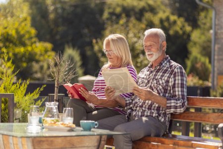 Foto de Feliz pareja de ancianos disfrutando de su tiempo juntos tomando un desayuno al aire libre en el patio trasero de su casa, hombre leyendo el periódico mientras que la mujer está leyendo un libro - Imagen libre de derechos