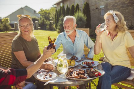 Photo for Group of cheerful elderly people making a toast while having lunch in the backyard, gathered around the table, eating, drinking and relaxing on a sunny summer day - Royalty Free Image
