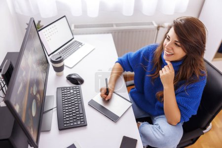 Photo for High angle view of female photographer sitting at her desk in home office retouching images using graphic pad and desktop computer while working remotely from home - Royalty Free Image