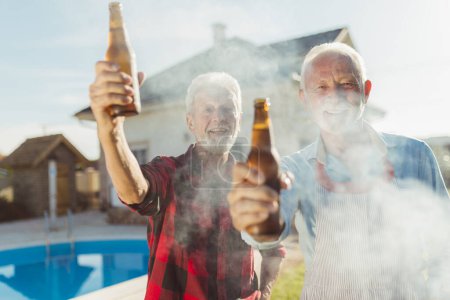 Foto de Dos hombres mayores haciendo un brindis con botellas de cerveza mientras asan carne a la parrilla en la fiesta de barbacoa del patio trasero junto a la piscina, relajándose al aire libre en el soleado día de verano - Imagen libre de derechos