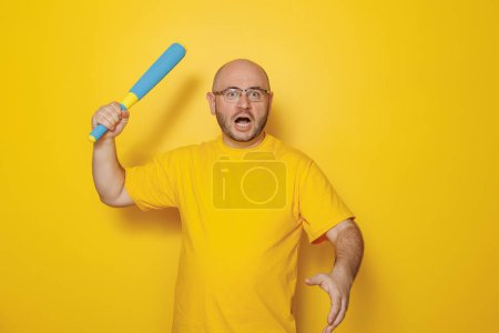 Téléchargez les photos : Mi-trentaine homme chauve en surpoids portant des lunettes tenant un jouet de baseball chauve-souris et faisant des visages sur fond jaune avec espace de copie - en image libre de droit