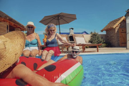 Foto de Dos hombres mayores enfriándose en la piscina mientras las mujeres beben cócteles, tomando el sol y relajándose junto a la piscina, divirtiéndose en las vacaciones de verano - Imagen libre de derechos