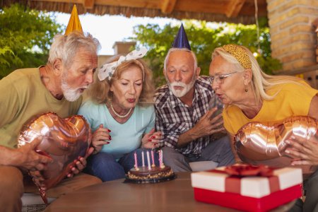 Téléchargez les photos : Groupe d'amis aînés joyeux s'amusant à une fête d'anniversaire, portant des chapeaux de fête, tenant des ballons et soufflant des bougies sur un gâteau d'anniversaire - en image libre de droit