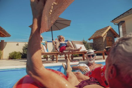 Foto de Dos parejas mayores disfrutando de sus vacaciones de verano juntas, mujeres tomando cócteles y tomando el sol en tumbonas junto a la piscina mientras los hombres nadan y se refrescan en la piscina - Imagen libre de derechos