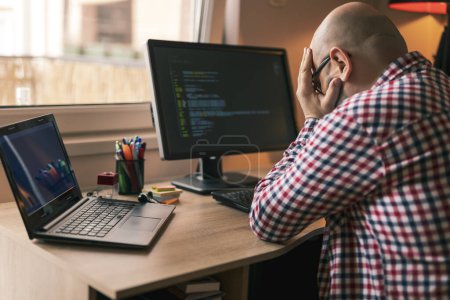 Photo for Software developer sitting at his desk while working from home, holding head in hands, stressed out because of code malfunction - Royalty Free Image