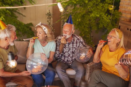 Photo for Group of cheerful senior friends having fun at a birthday party, holding balloons and blowing party whistles - Royalty Free Image