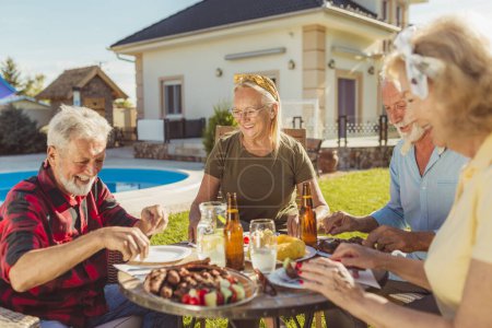 Photo for Group of senior people eating lunch in the backyard by the swimming pool, gathered around the table, eating, drinking and enjoying sunny summer day outdoors - Royalty Free Image