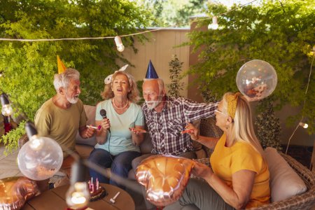 Foto de Grupo de personas mayores alegres teniendo una fiesta de cumpleaños para un amigo, divirtiéndose cantando karaoke mientras celebran - Imagen libre de derechos