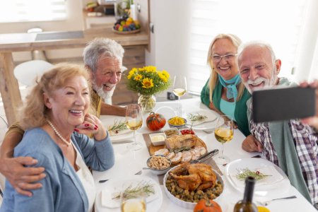 Foto de Personas mayores divirtiéndose mientras celebran Acción de Gracias juntas en casa durante la cena tradicional, tomando una selfie usando un teléfono inteligente - Imagen libre de derechos