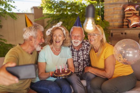 Photo for Group of cheerful senior people having fun celebrating friend's birthday, taking a selfie with birthday cake while at the party - Royalty Free Image