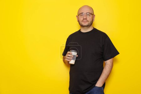 Téléchargez les photos : Portrait d'un homme chauve d'une trentaine d'années en surpoids portant un T-shirt noir et des lunettes tenant une tasse de café à emporter isolé sur fond jaune avec espace de copie - en image libre de droit