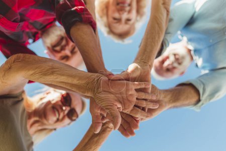 Photo for Low angle view of group of cheerful active senior people enjoying sunny summer day outdoors, gathered in circle, holding hands all together in the middle - Royalty Free Image