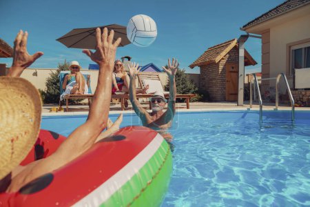 Photo for Two senior couples enjoying their summer vacation together, women drinking cocktails and sunbathing on sun beds by the pool while men are playing volleyball in the pool - Royalty Free Image