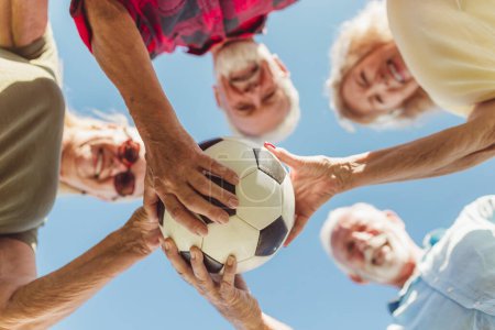 Foto de Vista de bajo ángulo de un grupo de alegres personas mayores activas que se divierten jugando al fútbol, se reunieron en círculo, sosteniendo una pelota de fútbol todos juntos antes del partido - Imagen libre de derechos