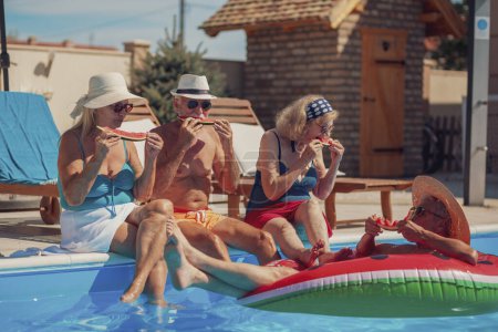Téléchargez les photos : Groupe de personnes âgées gaies assis au bord d'une piscine mangeant de la pastèque, prenant un bain de soleil et s'amusant à l'extérieur par une chaude journée d'été - en image libre de droit