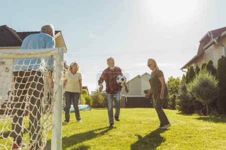 Photo for Group of active elderly people having fun playing football on the lawn in the backyard, enjoying sunny summer day outdoors, running after a ball - Royalty Free Image