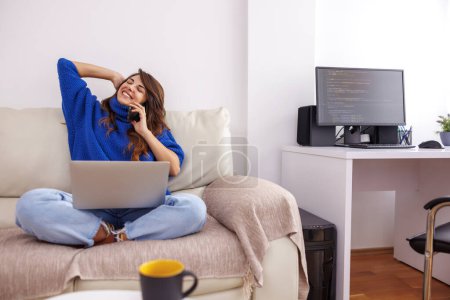 Foto de Hermosa mujer joven sentada en el sofá de la sala de estar que sostiene el ordenador portátil en su regazo y que tiene una llamada telefónica mientras trabaja de forma remota desde casa - Imagen libre de derechos