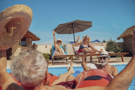 Photo for Two senior couples enjoying their summer vacation together, women drinking cocktails and sunbathing by the pool while men are swimming and cooling down in the pool - Royalty Free Image