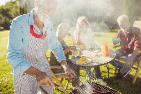 Photo for Handsome elderly man grilling meat at backyard barbecue party while his friends are sitting and relaxing in the background - Royalty Free Image