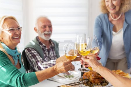 Photo for Group of senior people having fun while celebrating Thanksgiving together at home over traditional dinner, making a toast raising glasses of wine - Royalty Free Image