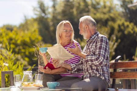 Foto de Feliz pareja de ancianos disfrutando de su tiempo juntos tomando un desayuno al aire libre en el patio trasero de su casa, hombre leyendo el periódico mientras que la mujer está leyendo un libro - Imagen libre de derechos