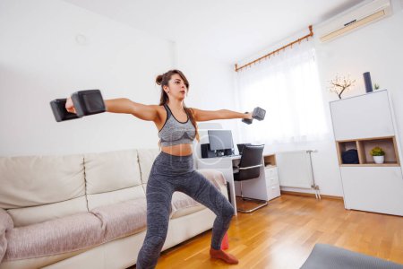 Photo for Active young woman in sportswear lifting weights while doing arm exercises at home - Royalty Free Image