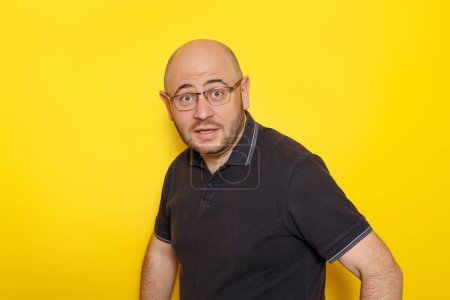 Photo for Cheerful funny mid 30s bald overweight man wearing black T-shirt and glasses making faces isolated on yellow colored background - Royalty Free Image