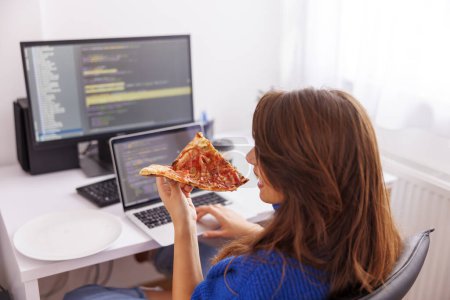 Téléchargez les photos : Programmeuse assise à son bureau à la maison, prenant une pause tout en travaillant à distance de la maison en buvant du café et en mangeant de la pizza - en image libre de droit