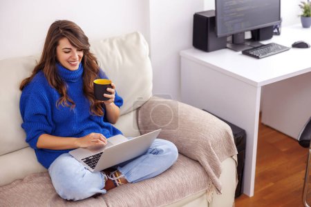 Téléchargez les photos : Jeune femme assise sur le canapé du salon, tenant un ordinateur portable sur ses genoux et prenant une pause café tout en travaillant à la maison - en image libre de droit