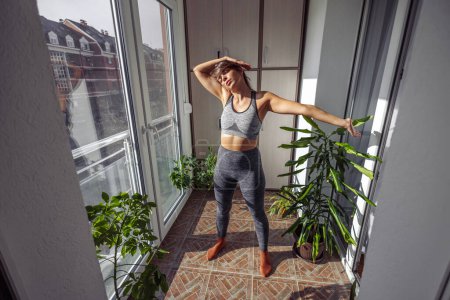 Photo for Young woman in sportswear stretching while working out at home in the morning - Royalty Free Image