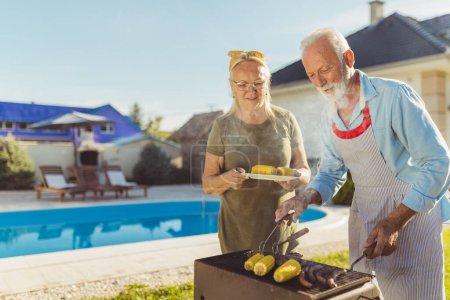 Téléchargez les photos : Couple âgé ayant une fête barbecue arrière-cour, griller de la viande et du maïs, et avoir du plaisir à passer du temps libre à l'extérieur - en image libre de droit