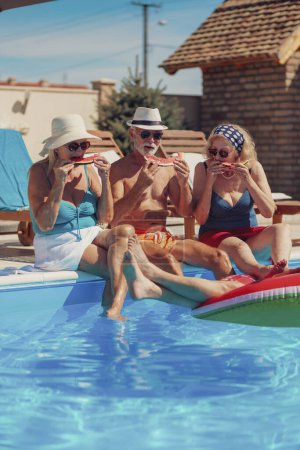 Photo for Group of cheerful senior people sitting at the edge of a swimming pool eating watermelon slices, sunbathing and having fun outdoors on a hot summer day - Royalty Free Image