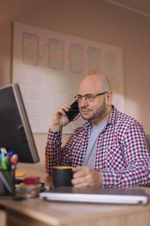 Téléchargez les photos : Homme ayant une conversation téléphonique et buvant du café tout en travaillant dans un bureau - en image libre de droit