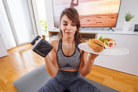 Foto de Mujer en ropa deportiva sosteniendo una mancuerna en una mano y sándwich en un plato en el otro - elección entre estilo de vida saludable y entrenamiento y comida rápida - Imagen libre de derechos