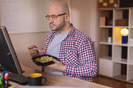 Téléchargez les photos : Homme assis à son bureau dans un bureau, prenant une pause déjeuner, mangeant des pâtes à emporter et buvant du café - en image libre de droit