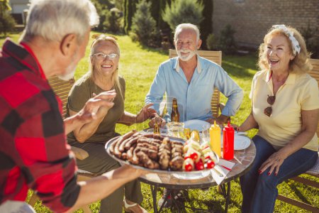 Foto de Grupo de alegres amigos mayores almorzando al aire libre en el patio trasero, reunidos alrededor de la mesa, anfitriones llevando comida en una bandeja, ofreciéndosela a los invitados - Imagen libre de derechos