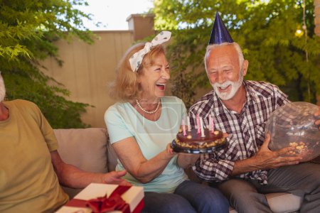 Photo for Group of cheerful senior friends having fun at a birthday party, host of the party holding a birthday cake after making a wish and blowing out candles - Royalty Free Image