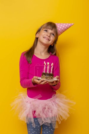Téléchargez les photos : Belle petite fille portant un chapeau d'anniversaire, tenant un gâteau d'anniversaire avec des bougies allumées dessus, faisant un souhait et soufflant des bougies, isolé sur fond jaune - en image libre de droit