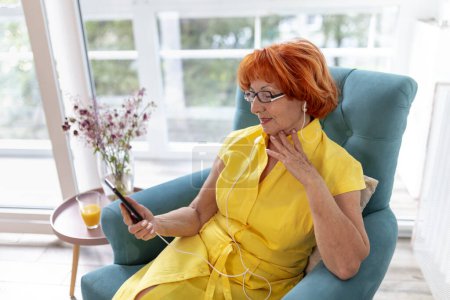 Téléchargez les photos : Femme âgée se détendre à la maison, profiter de son temps libre, écouter la musique et choisir une chanson à partir d'une liste de lecture de téléphone intelligent - en image libre de droit