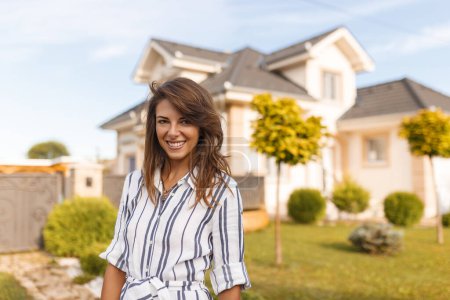 Foto de Hermosa joven mujer de pie frente a su nueva casa feliz después de comprar una nueva propiedad - Imagen libre de derechos