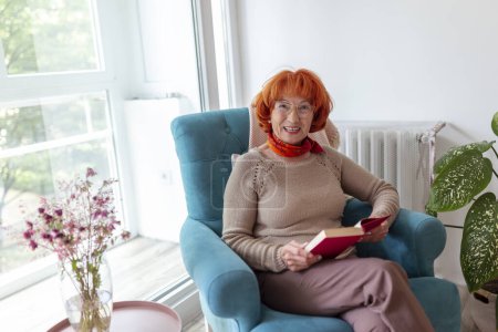 Foto de Mujer mayor sentada en un sillón junto a la ventana, leyendo un libro y relajándose en casa - Imagen libre de derechos