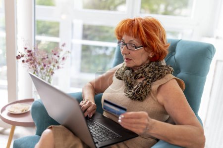 Téléchargez les photos : Belle femme âgée assise dans un fauteuil, relaxante à la maison, utilisant un ordinateur portable, payant en ligne à l'aide d'une carte de crédit ; femme âgée faisant du shopping sur Internet - en image libre de droit