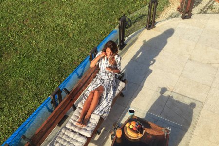 Téléchargez les photos : Vue grand angle d'une belle jeune femme se relaxant dans la cour de sa maison et en utilisant un téléphone intelligent - en image libre de droit